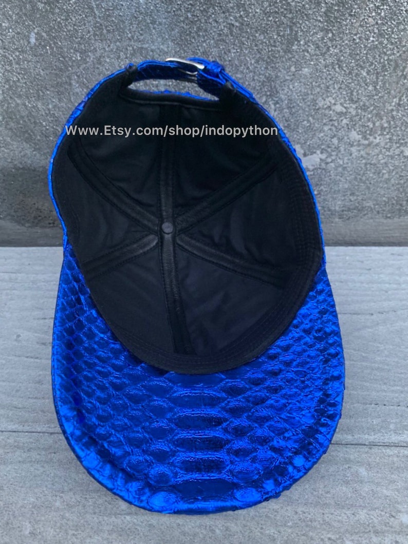 May include: A blue baseball cap with a snakeskin pattern. The inside of the cap is black with a white stitching detail. The cap has a black adjustable strap with a silver buckle. The text "www.Etsy.com/shop/indopython" is printed on the inside of the cap.
