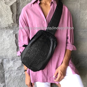 Könnte beinhalten: Eine schwarze Leder-Umhängetasche mit Totenkopf-Design. Die Tasche hat einen langen Riemen und ist aus Pythonleder gefertigt.