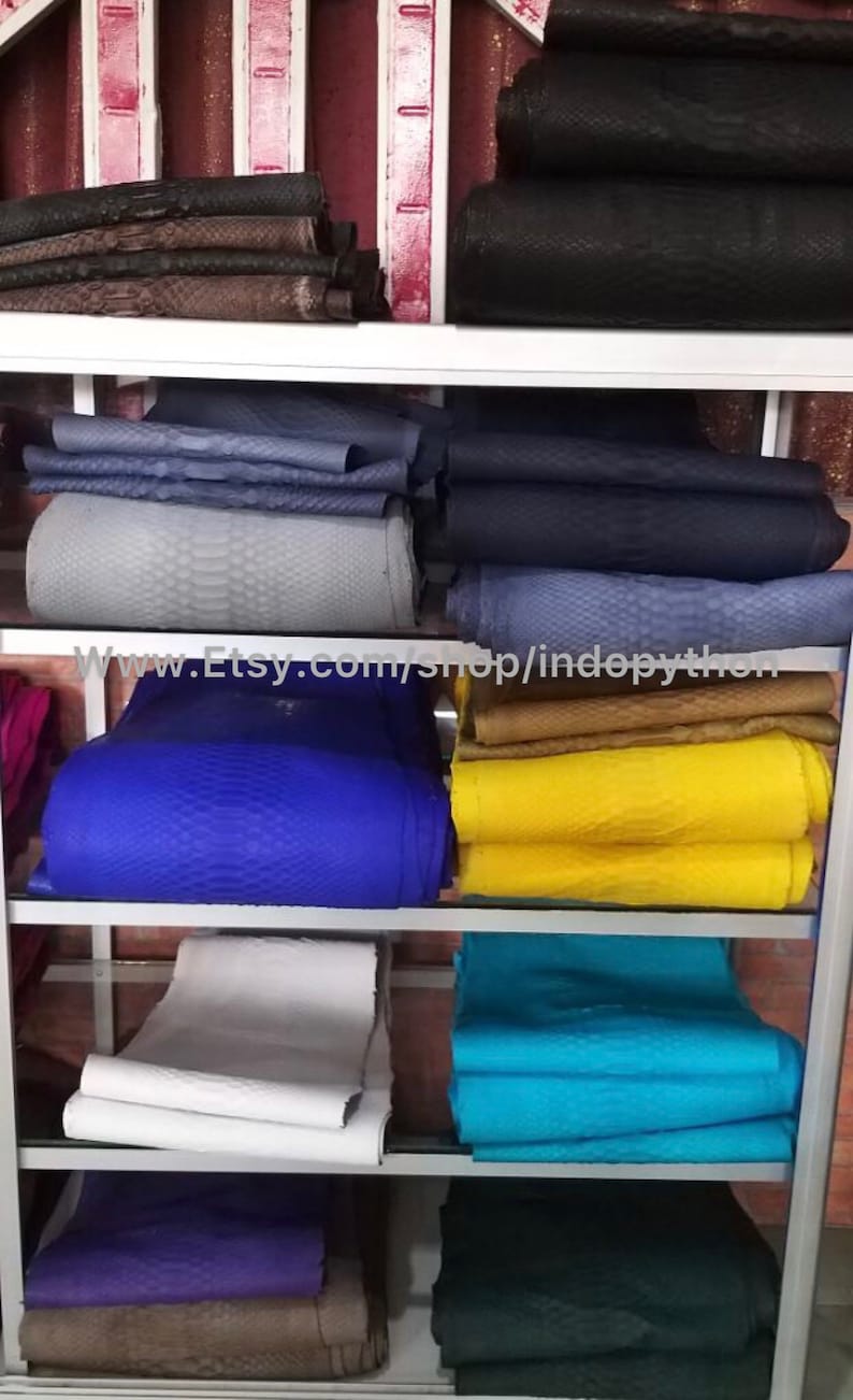 Może przedstawiać: P&oacute;łka z ekspozycją r&oacute;żnych kolor&oacute;w sk&oacute;ry pytona. Sk&oacute;ra jest ułożona w stosy na białej p&oacute;łce z metalową ramą. Sk&oacute;ra jest dostępna w kolorach takich jak niebieski, ż&oacute;łty, biały i zielony. Adres strony internetowej www.Etsy.com/shop/indopython jest widoczny na p&oacute;łce.