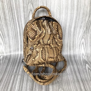 May include: A small, brown and tan backpack with a snakeskin pattern. The backpack has a top handle, a zippered closure, and adjustable straps. The bag is made of a textured material and has a small, round shape. The text "com/shop/indopy" is visible.
