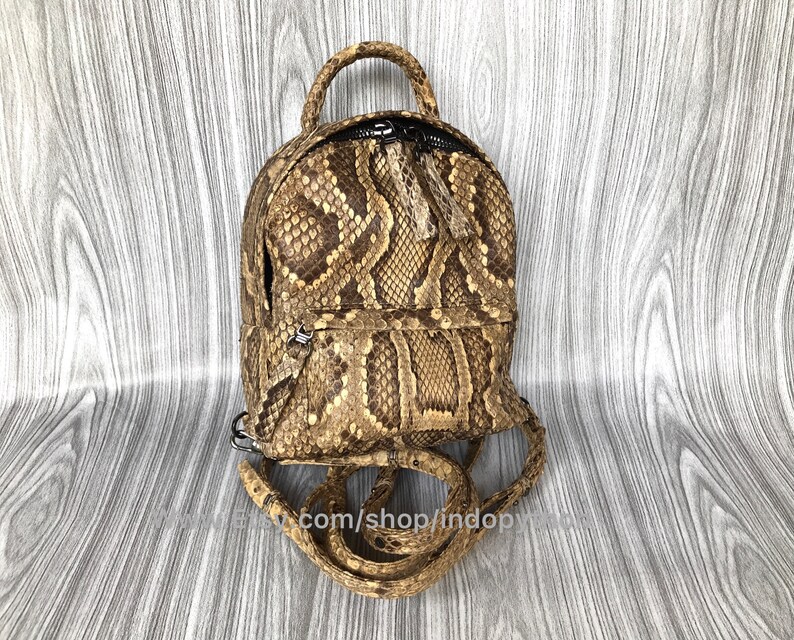 May include: A small, brown and tan backpack with a snakeskin pattern. The backpack has a top handle, a zippered closure, and adjustable straps. The bag is made of a textured material and has a small, round shape. The text "com/shop/indopy" is visible.