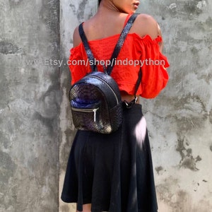 Pu&ograve; includere: Zaino nero e blu iridescente con tasca frontale con cerniera e spallacci regolabili. Lo zaino presenta un motivo strutturato e scaglioso. L'URL del negozio Etsy &egrave; visibile nell'immagine.