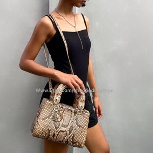Puede incluir: Un bolso de mano de piel de serpiente marrón y beige con cierre dorado y correa de cadena dorada. El bolso lo lleva una persona que lleva un top negro sin mangas.