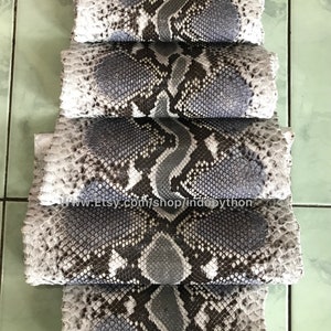 May include: A stack of gray and black python skins with a white background. The skins are arranged in a staggered pattern, with the top skin being the largest and the bottom skin being the smallest. The skins are all smooth and have a natural, textured pattern.
