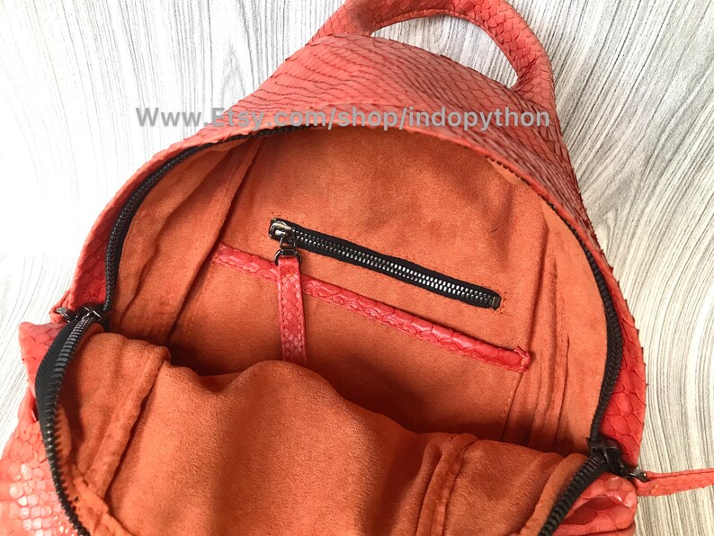 Pu&ograve; includere: L'interno di uno zaino in pelle di serpente arancione brillante con una tasca con cerniera e una fodera arancione morbida. Lo zaino ha un manico superiore e una chiusura a cerniera.