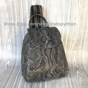 K&ouml;nnte beinhalten: Ein grauer Schlangenleder-Rucksack mit Rei&szlig;verschluss. Der Rucksack hat eine Klappe mit Schnallenverschluss. Der Rucksack befindet sich auf einer Holzoberfl&auml;che.