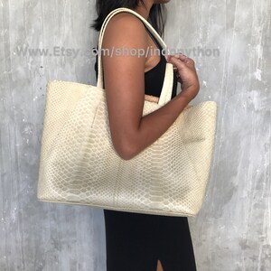 May include: A large, off-white tote bag made of python skin. The bag has a wide, open top and two long handles.