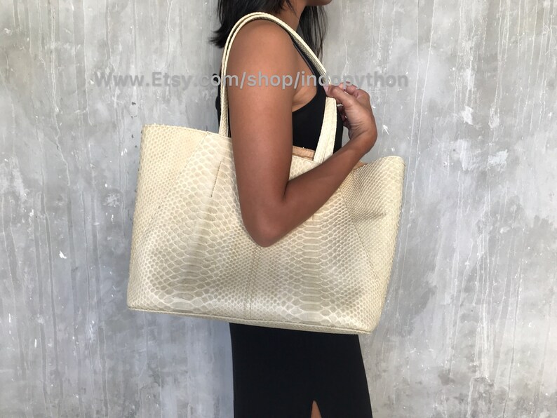 May include: A large, off-white tote bag made of python skin. The bag has a wide, open top and two long handles.