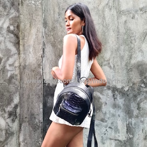 Może przedstawiać: Czarny plecak ze skóry węża z srebrnymi okuciami i zamkiem błyskawicznym. Plecak nosi osoba w białej sukience.