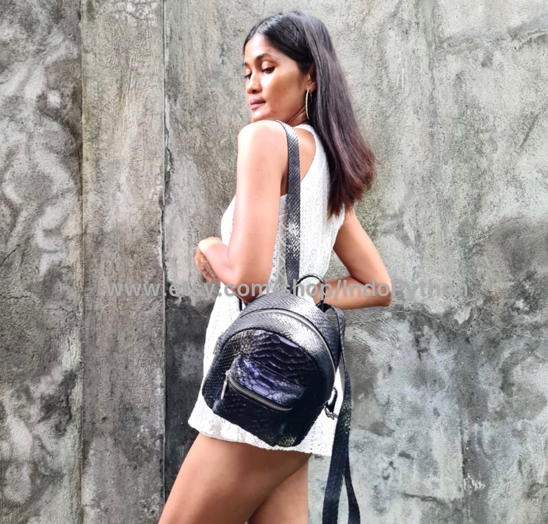 May include: A black snakeskin backpack with silver hardware and a zipper closure. The backpack is worn by a person in a white dress.
