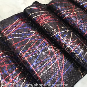 May include: Close-up of several pieces of black snakeskin leather with a colourful abstract design. The design features red, blue, and white lines over the black scales. The leather appears to be rolled or folded.