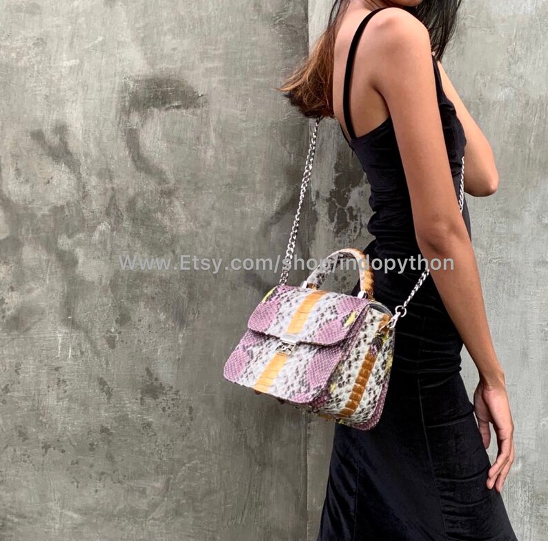 May include: A small, pink, white, and orange snakeskin handbag with a silver chain strap and a top handle. The bag is worn by a person wearing a black dress.