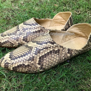 Op de afbeelding: Een paar beige en bruine slangenleren slippers met een spitse neus. De slippers liggen op een bed van groen gras.
