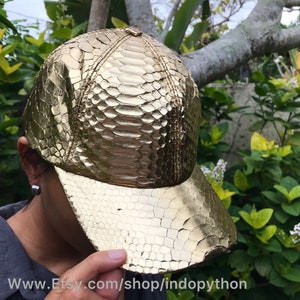 Può includere: Un berretto da baseball dorato in pelle di pitone. Il berretto ha una visiera curva e una corona strutturata.