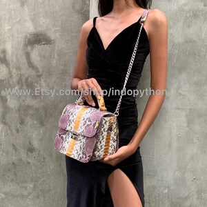 May include: A small, rectangular handbag with a snakeskin pattern in shades of purple, yellow, and white. The bag has a top handle and a chain strap. The bag is being held by a person wearing a black dress.