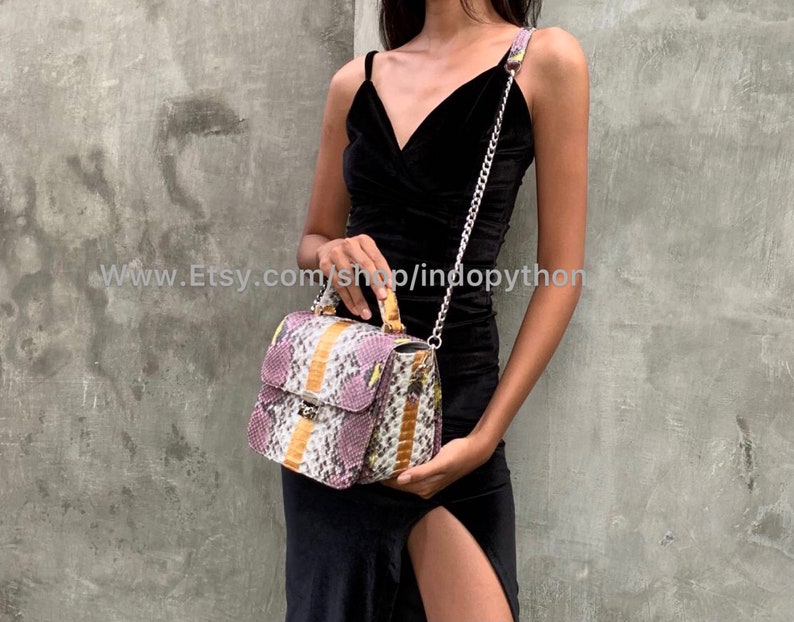 May include: A small, rectangular handbag with a snakeskin pattern in shades of purple, yellow, and white. The bag has a top handle and a chain strap. The bag is being held by a person wearing a black dress.