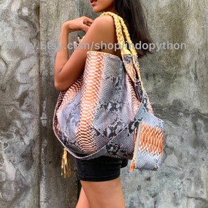 Op de afbeelding: Een grote, grijze, oranje en witte slangenleren tote bag met een gele gevlochten band en een kleinere bijpassende slangenleren tas. De tas heeft een kwast aan de zijkant.