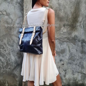 May include: A black leather backpack with a snakeskin pattern and beige straps. The backpack has two buckles on the front and is worn by a person.