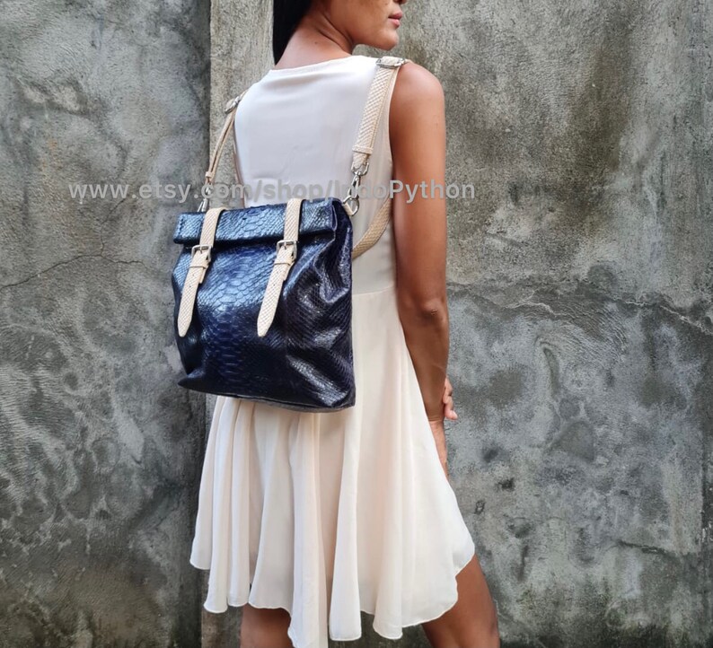 May include: A black leather backpack with a snakeskin pattern and beige straps. The backpack has two buckles on the front and is worn by a person.