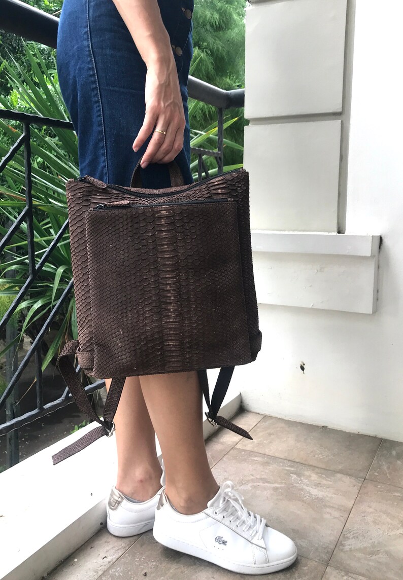 K&ouml;nnte beinhalten: Ein brauner Rucksack aus Schlangenleder mit verstellbaren Tr&auml;gern. Der Rucksack hat eine Rei&szlig;verschlusstasche vorne und einen Tragegriff oben.