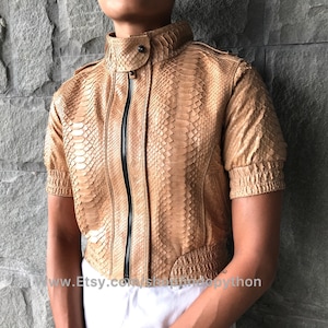 Puede incluir: Chaqueta corta color beige con estampado de piel de serpiente y mangas cortas. Presenta cuello alto, cremallera negra y botones en los hombros. Las mangas están fruncidas en los puños. La chaqueta es de material texturizado.