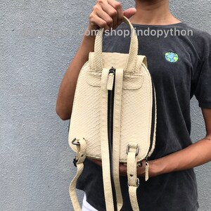 May include: A small, off-white leather backpack with a zipper closure and adjustable straps. The backpack has a textured, snakeskin-like pattern.