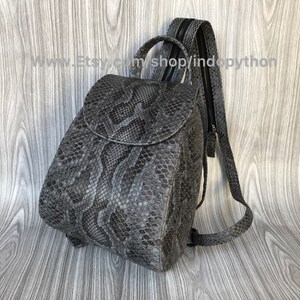 K&ouml;nnte beinhalten: Ein kleiner grauer Schlangenleder-Rucksack mit Rei&szlig;verschluss und verstellbaren Tr&auml;gern. Der Rucksack hat eine abgerundete Form und einen Klappenverschluss.