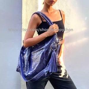 May include: A blue python leather tote bag with a gold ring and tassel. The bag has a long strap and is worn over the shoulder.