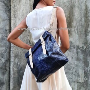 May include: A dark blue leather satchel with a white strap and buckle accents. The bag has a textured, snakeskin-like pattern.
