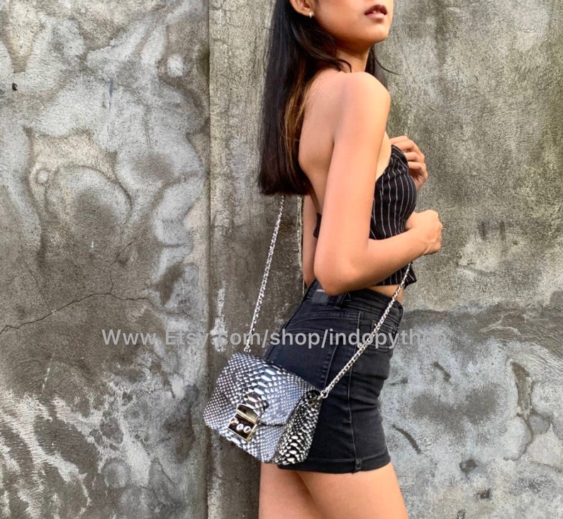 May include: A small black and white snakeskin crossbody bag with a silver chain strap and a silver lock closure. The bag is worn by a person wearing a black and white striped top and black denim shorts.