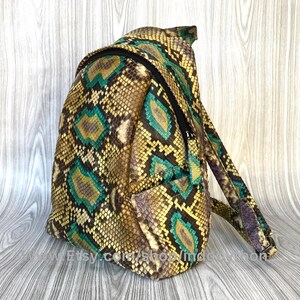Peut inclure: Un petit sac &agrave; dos en peau de serpent vert et jaune. Le sac &agrave; dos a une poche zipp&eacute;e &agrave; l'avant et une sangle qui passe par-dessus l'&eacute;paule.