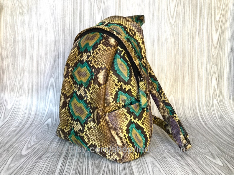 Peut inclure: Un petit sac &agrave; dos en peau de serpent vert et jaune. Le sac &agrave; dos a une poche zipp&eacute;e &agrave; l'avant et une sangle qui passe par-dessus l'&eacute;paule.