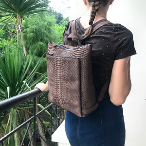 K&ouml;nnte beinhalten: Ein brauner Schlangenleder-Rucksack mit Rei&szlig;verschluss. Der Rucksack wird von einer Person mit einem Zopf getragen.