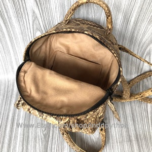 May include: Open, tan and brown snakeskin backpack with a black zipper and tan interior. The bag has a top handle and adjustable straps. The interior features a small pocket. The bag is open, showing the inside.