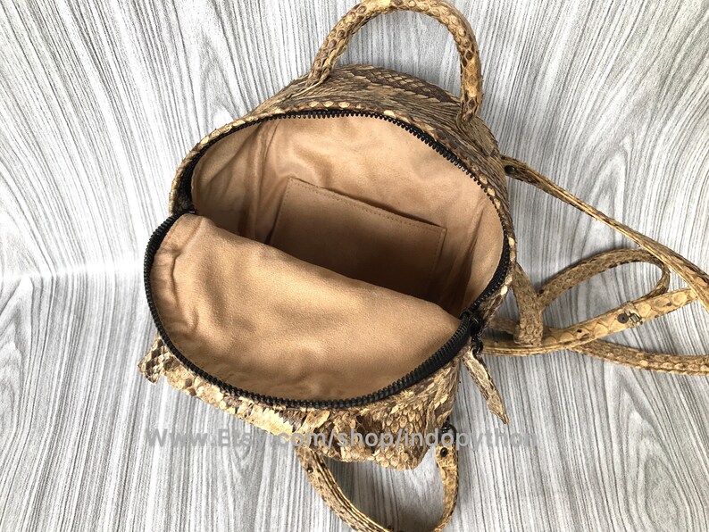 May include: Open, tan and brown snakeskin backpack with a black zipper and tan interior. The bag has a top handle and adjustable straps. The interior features a small pocket. The bag is open, showing the inside.