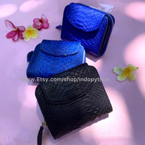 K&ouml;nnte beinhalten: Drei kleine Rei&szlig;verschlussportemonnaies aus Pythonleder. Die Portemonnaies sind in Blaut&ouml;nen und Schwarz gehalten. Die Portemonnaies sind auf einer rosa Oberfl&auml;che mit wei&szlig;en Blumen angeordnet.