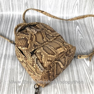 May include: A brown and tan snakeskin backpack with adjustable straps. The bag has a rounded shape with a front pocket and a zipper closure. The snakeskin pattern is visible across the entire bag, including the straps.