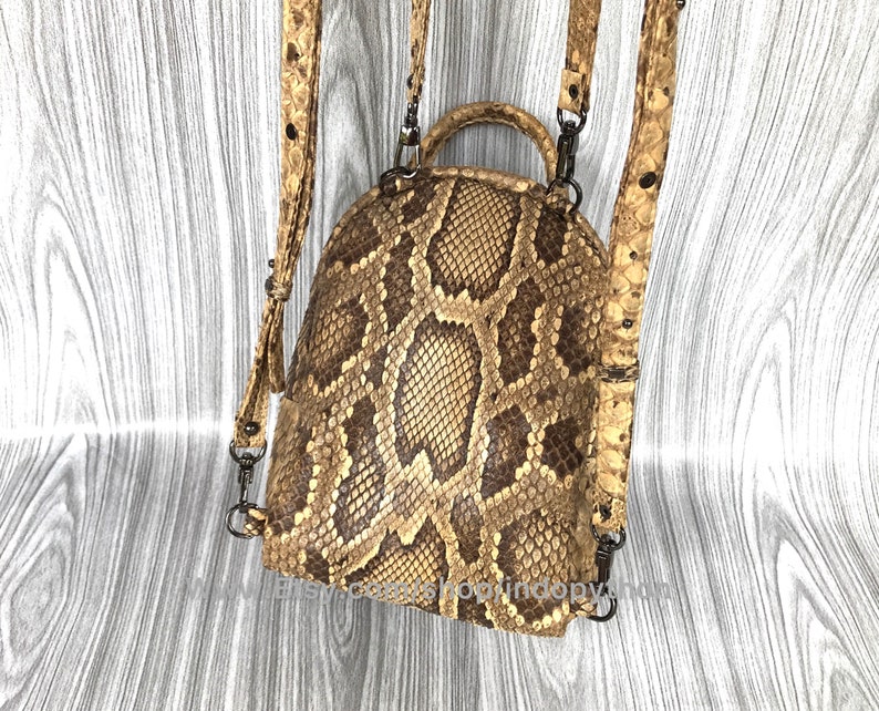 May include: A brown and tan snakeskin backpack with adjustable straps and a top handle. The backpack has a rounded shape and features a repeating pattern of scales. The straps and handle are a lighter shade of tan with black hardware.