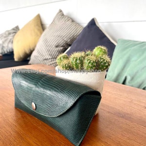 Op de afbeelding: Een donkergroene, getextureerde leren brillenkoker met een drukknoopsluiting. De koker staat op een houten tafel naast een kleine cactus in een pot. De achtergrond bevat kleurrijke kussens.