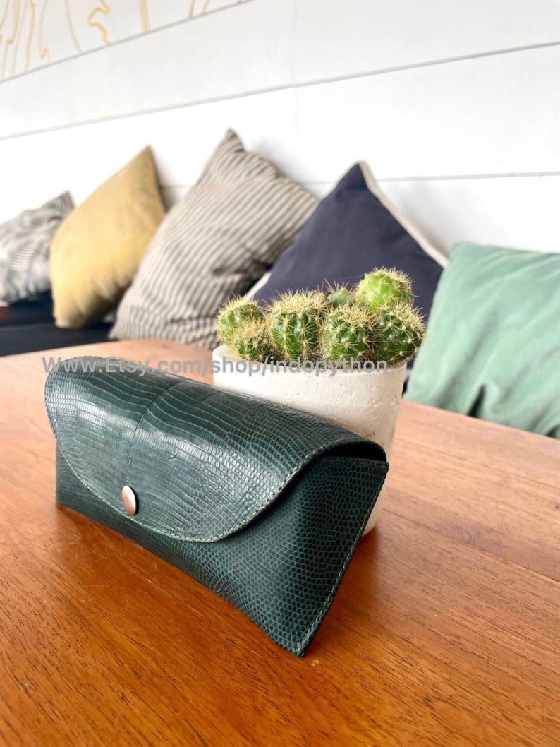 Op de afbeelding: Een donkergroene, getextureerde leren brillenkoker met een drukknoopsluiting. De koker staat op een houten tafel naast een kleine cactus in een pot. De achtergrond bevat kleurrijke kussens.
