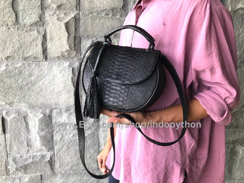 May include: A black leather handbag with a snakeskin pattern and a tassel. The bag has a top handle and a long strap for carrying on the shoulder.