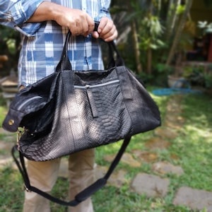 Puede incluir: Una bolsa de viaje de cuero negro con un patrón de piel de serpiente. La bolsa tiene una cremallera y una correa de hombro larga.