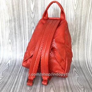 Pu&ograve; includere: Uno zaino arancione brillante in pelle di serpente con spalline regolabili. Lo zaino ha una tasca con cerniera sul davanti e un manico superiore.