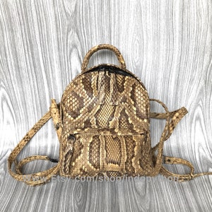 May include: A small, round backpack made of snakeskin. The bag features a top handle, adjustable straps, and a front zippered pocket. The snakeskin pattern is a mix of tan, brown, and black.