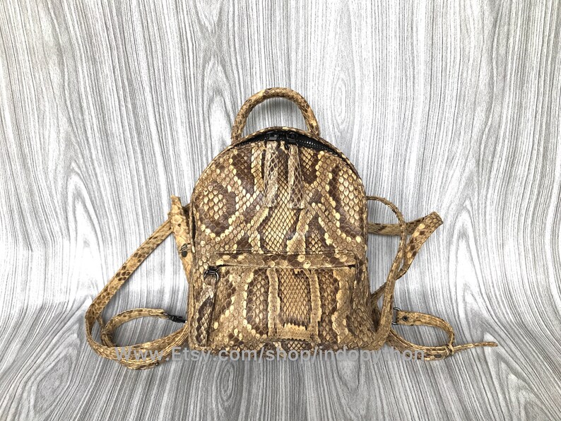 May include: A small, round backpack made of snakeskin. The bag features a top handle, adjustable straps, and a front zippered pocket. The snakeskin pattern is a mix of tan, brown, and black.