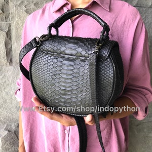 May include: A black round handbag made of python skin with a top handle and a long shoulder strap. The bag has a flap closure with a silver buckle.