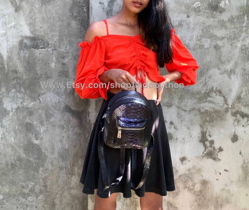 Pu&ograve; includere: Un piccolo zaino nero con un motivo strutturato simile alla pelle di serpente. La borsa ha una tasca frontale con cerniera e spallacci regolabili. L'URL del negozio Etsy, www.Etsy.com/shop/indopython, &egrave; visibile.