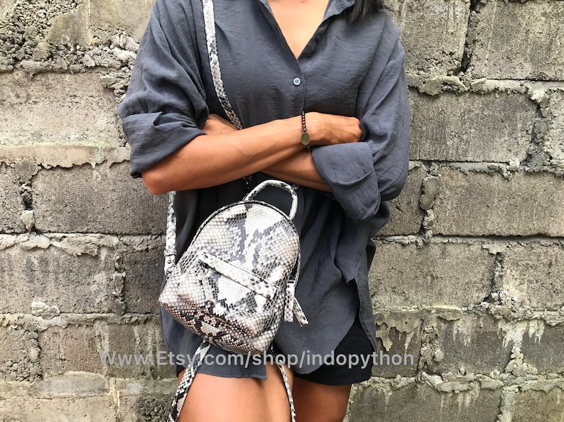 K&ouml;nnte beinhalten: Ein kleiner, wei&szlig;er und schwarzer Schlangenleder-Rucksack mit Rei&szlig;verschluss und silberner Kettenriemen. Der Rucksack wird von einer Person getragen, die ein graues Hemd und schwarze Shorts tr&auml;gt. Der Text "www.Etsy.com/shop/indopython" ist am unteren Rand des Bildes sichtbar.