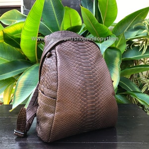 May include: A brown leather backpack with a snakeskin pattern. The backpack has a flap closure, adjustable straps, and a side pocket. The text "www. indopython.com/shop/indopython" is visible on the image.