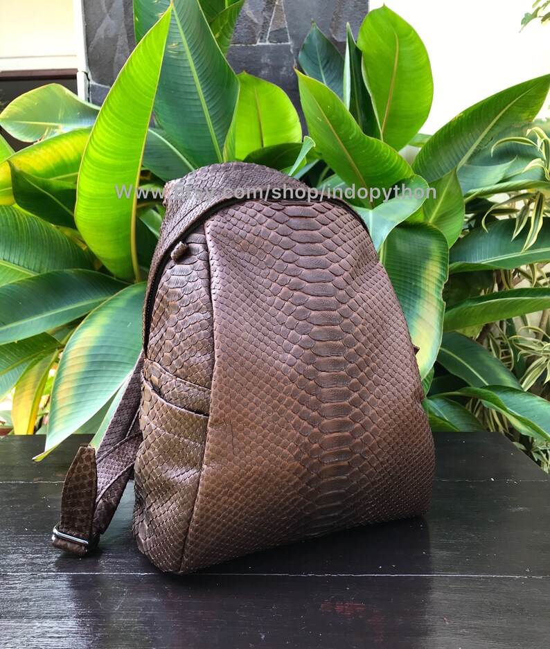 May include: A brown leather backpack with a snakeskin pattern. The backpack has a flap closure, adjustable straps, and a side pocket. The text "www. indopython.com/shop/indopython" is visible on the image.